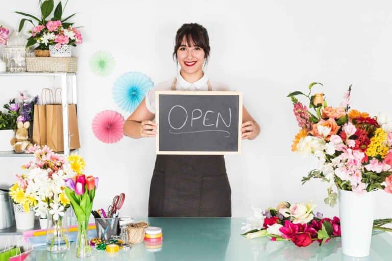 A woman florist holding a shop open board in a flower shop.